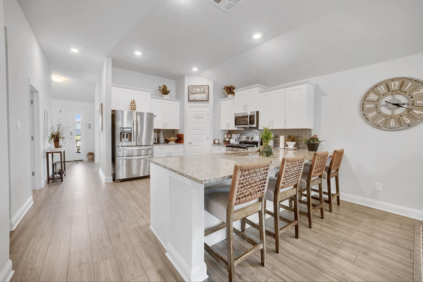 Open kitchen with granite island, four woven barstools, and a view down the entry hallway